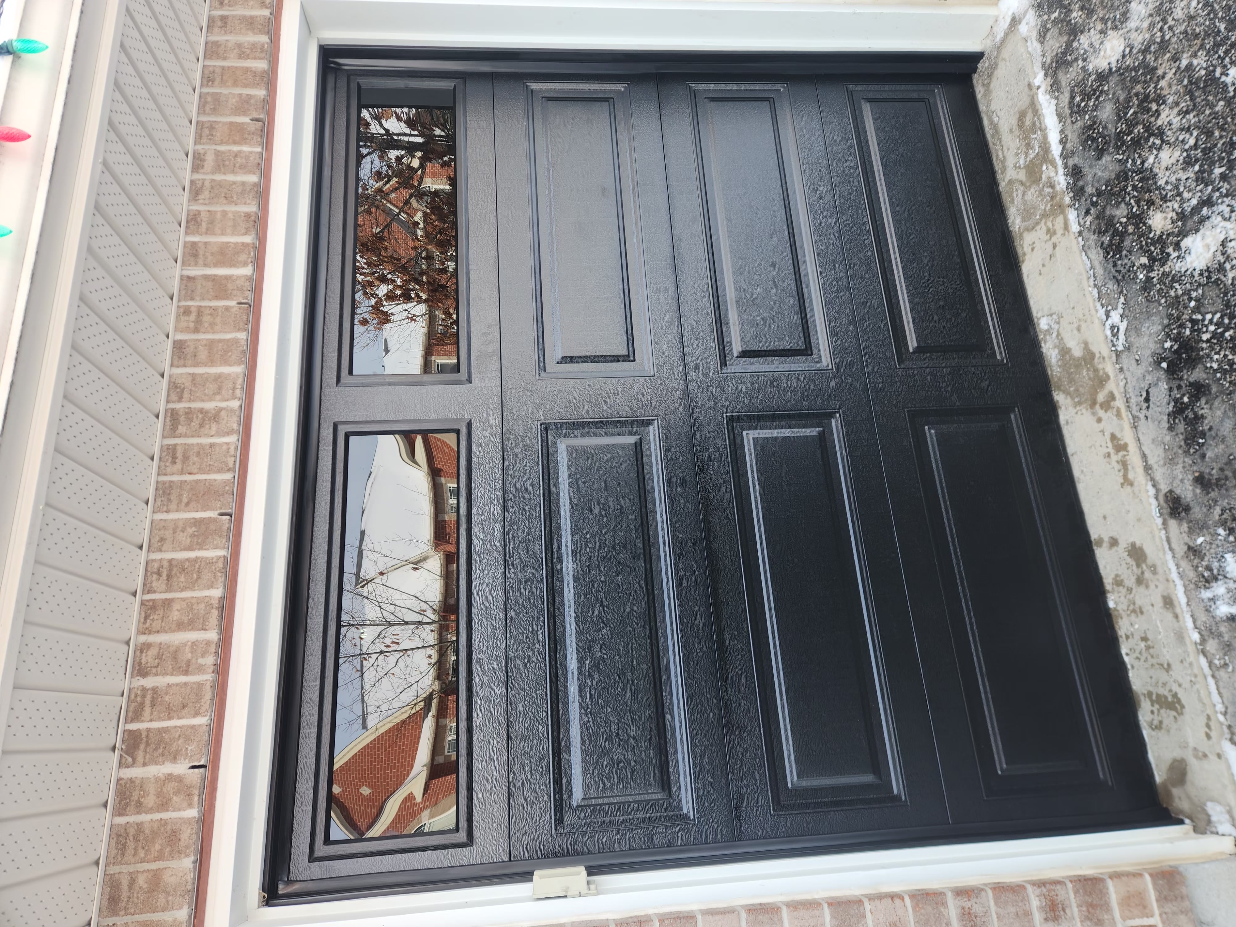 New black raised-panel single-car garage door with two windows installed on brick Stittsville home